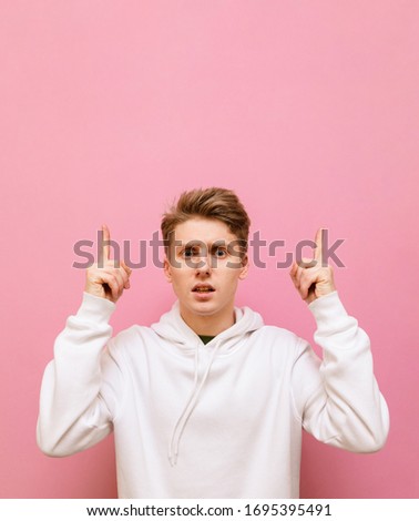 Surprised guy in white hoodie isolated on pink background, looks into camera and shows thumbs up on copy space. Vertical portrait of shocked young man pointing finger at empty space