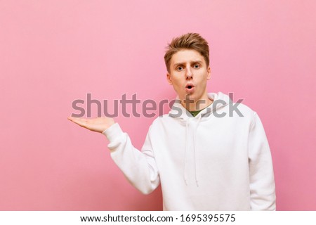 Surprised guy in white hoodie stands on pink background, holds copy space in his hand and looks into the camera. Shocked young man holding his hand aside and looking away, isolated.