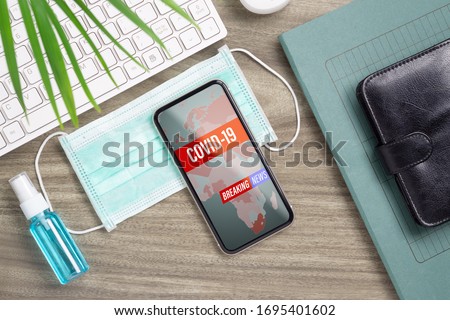 Coronavirus or Covid-19 outbreak Breaking News update background concept. Mockup mobile phone with facial masks and Alcohol Mini Hand Sanitizer Spray on office table workspace. Royalty-Free Stock Photo #1695401602