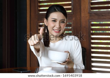 Young asian woman sit at rustic cafe holding soup noodle bowl hand finger thumbs up good sign look front at camera focus on face