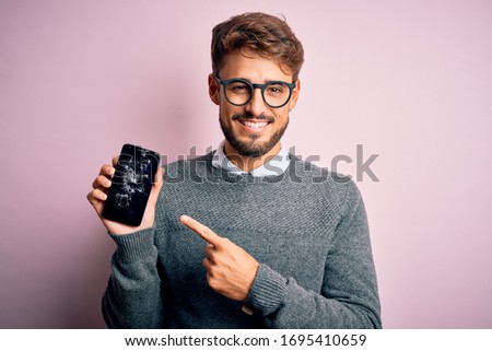 Young man with beard wearing glasses holding broken and craked smartphone very happy pointing with hand and finger