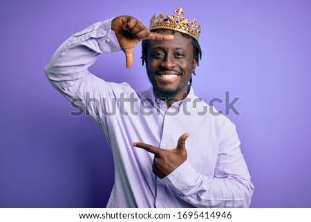 Young african american man wearing golden crown of king over isolated purple background smiling making frame with hands and fingers with happy face. Creativity and photography concept.