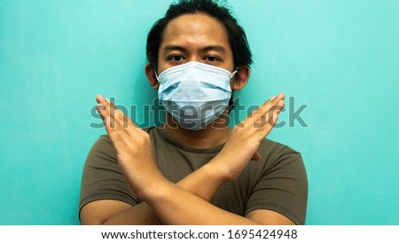 A portrait of a young Malay man wearing 3 layer surgical face mask showing crossing hand indicated NO and Stop spreading virus on isolated blue background. Selective focused.