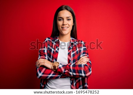 Young beautiful brunette woman wearing casual shirt standing over isolated red background happy face smiling with crossed arms looking at the camera. Positive person.