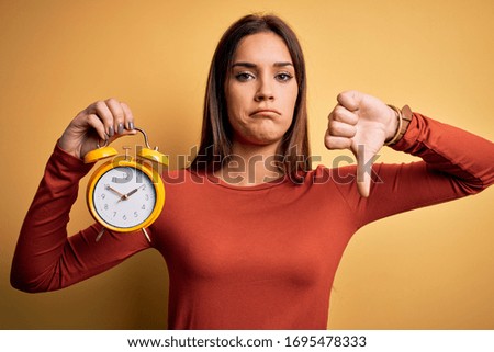 Young beautiful brunette woman holding alarm clock over isolated yellow background with angry face, negative sign showing dislike with thumbs down, rejection concept