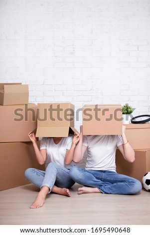 moving day - young couple covering their faces with cardboard boxes and copy space over white brick wall background