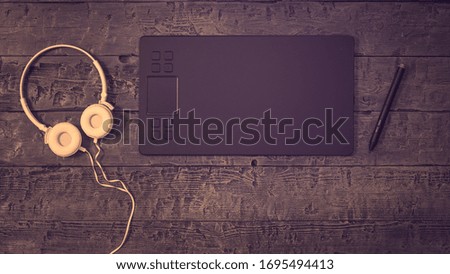 A tinted image of a graphics tablet with white headphones on a wooden background. A device for working in image editors.