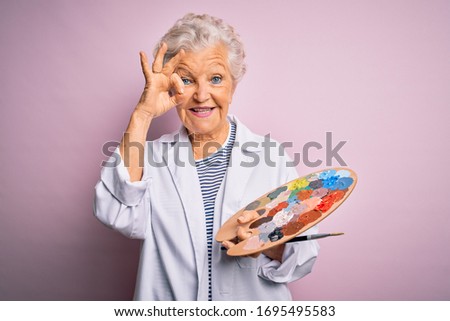 Senior beautiful grey-haired artist woman painting using brush and palette over pink background doing ok sign with fingers, excellent symbol