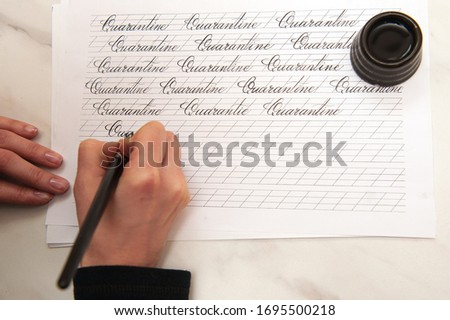 Female hands on distance learning write the word quarantine in calligraphy copybooks. Pen with black holder in black ink on white paper .standing black inkwell