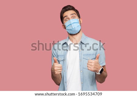 Portrait of young man with surgical medical mask in blue casual style shirt standing, thumbs up and looking at camera with surprised happy face. indoor studio shot, isolated on pink background. Royalty-Free Stock Photo #1695534709