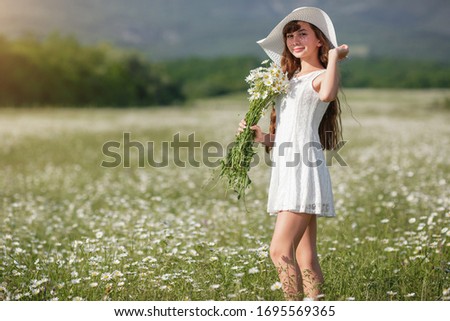 Beautiful cute teen girl on a walk in a daisy field, in a white dress and white hat. Idea and concept of happy growing up, health and allergy, skin and hair care.