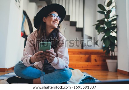 Happy woman enjoying sunny day relaxing at home talking online video chat with friends via cellphone sitting on floor in living room, Positive young hipster girl using smartphone typing text message