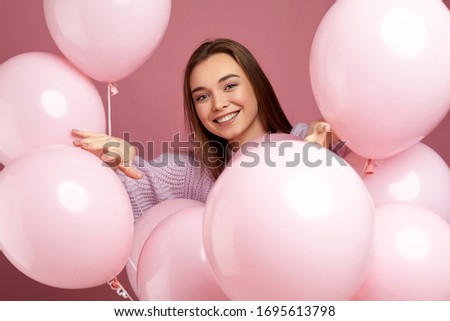 Smiling pleased girl posing with pastel pink air balloons on pink background. Beautiful happy young woman on a birthday holiday. close-up