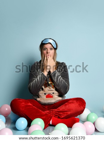 Teenage girl in gray reindeer plush pajamas, red socks, sleep mask. Covered mouth with palms, surprised, sitting on floor, colorful balloons, blue background. Christmas holidays. Copy space. Close up