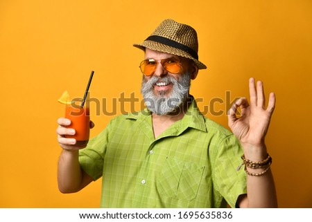 Bearded grandpa in hat, green shirt, sunglasses. He smiling, showing okay sign, holding glass of fresh squeezed juice with slice of lemon and tube, posing on orange background. Close up, copy space