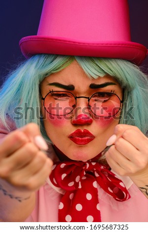 Close-up portrait of a clown girl with makeup, blue hair, in a pink hat, pink dress and glasses. A clown shows different human emotions. April Fools Day concept. Circus performance with a clown.