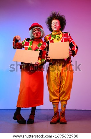 Two clowns a man and a woman with makeup in bright colored costumes are holding a blank cardboard for inscription. April Fools Day concept. Circus performance with a clown.