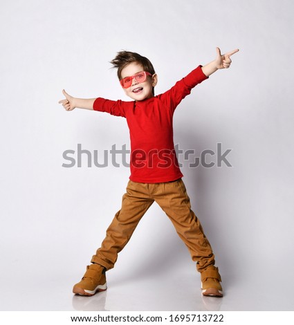 Little boy kid child, stylish hairstyle in sunglasses, red jumper smiling spread hands, pointing fingers showing tongue posing on white background. Childhood fashion advertising. Close up, copy space