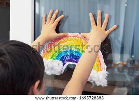 Kid putting painting colouful rainbow with cotton wool on white paper in the window during Covid-19 quarantine at home.Stay at home Social media campaign for coronavirus prevention concept