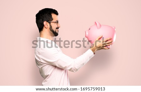Handsome man with beard holding a piggybank over isolated pink background