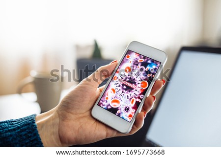 Closeup of a mobile cell phone of a young woman staying home and leadling business from distance on a freelance basis. Laptop, cup and flower. Coronavirus sign on the screen