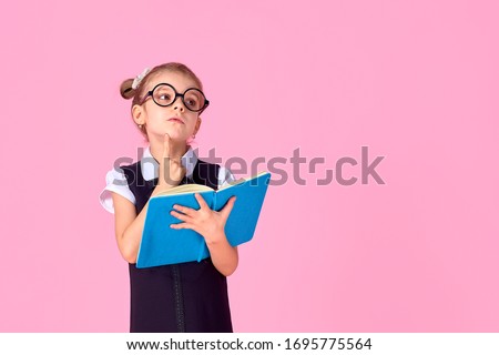 primary school girl in uniform, round glasses without lenses holds a notebook in her hands with a thoughtful emotion on her face, posing on a pink background in the studio.