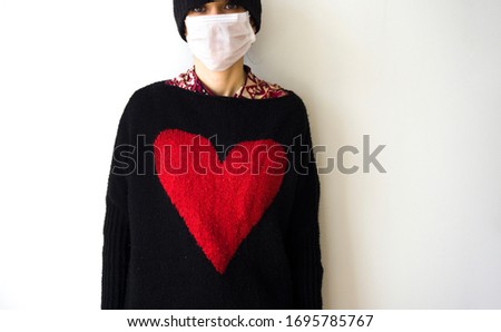 Young woman protecting herself from Corona virus (covid 19) with a mask on white background. Girl wearing a mask for potential winter colds. Female looking at camera, posing with red heart sign, shape