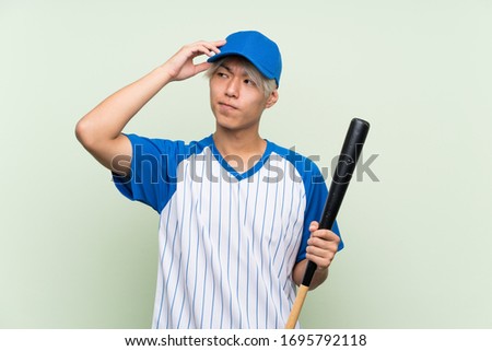 Young asian man playing baseball over isolated green background having doubts and with confuse face expression