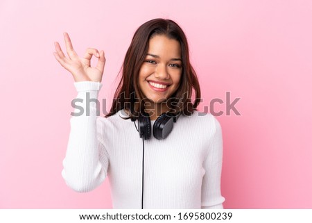 Young woman with earphones over isolated pink wall showing ok sign with fingers