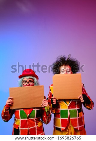 Two clowns a man and a woman with makeup in bright colored costumes are holding a blank cardboard for inscription. April Fools Day concept. Circus performance with a clown.