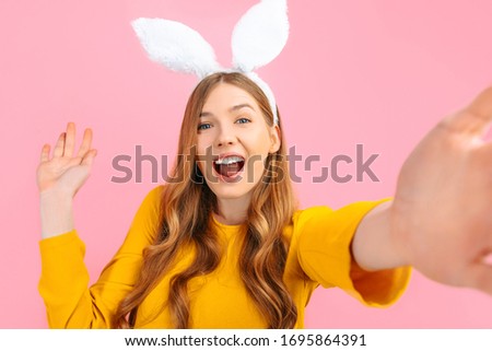 happy Easter. Smiling stylish woman in Easter Bunny ears, taking a selfie on her phone, on an isolated pink background
