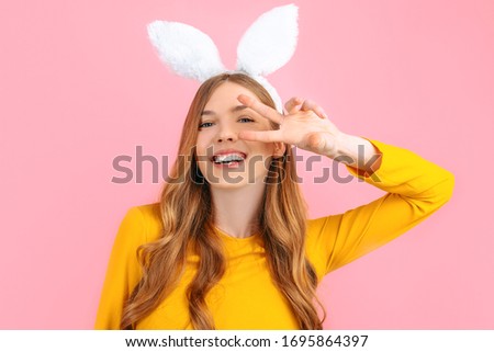 happy Easter. Smiling stylish woman in Easter Bunny ears, shows peace gesture, two fingers, on isolated pink background