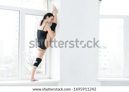 Full length portrait of a sporty slim girl doing splits standing on the windowsill in studio