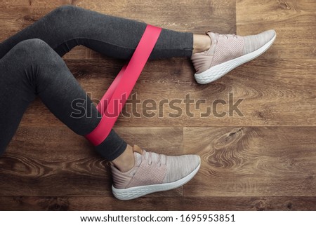 Female legs in sports leggings and sneakers doing exercises with fitness elastic on a wooden floor. Fitness workout at home. Top view