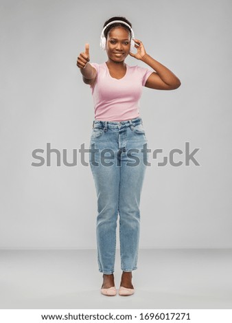 people, technology and audio equipment concept - happy african american young woman in headphones listening to music and showing thumbs up over grey background