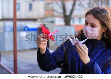 Pink and white origami paper cranes in the hands of a teenage girl in a medical respiratory mask. The concept of hope, faith, happy future, peace during  coronavirus epidemic, covid-19 pandemic. 