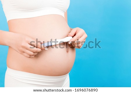 Close up portrait of pregnant woman in white underwear holding positive pregnancy test against her belly at blue background. Healthy pregnancy. Copy space.