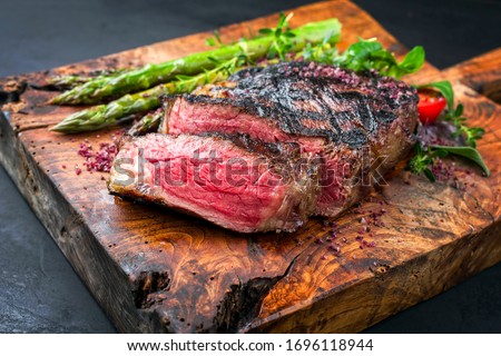 Barbecue dry aged wagyu entrecote beef steak with lettuce and green asparagus as closeup on an old rustic wooden cutting board  Royalty-Free Stock Photo #1696118944