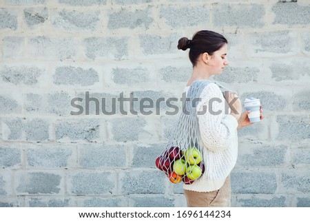 Young woman in light clothes with eco bag with apples and reusable coffee mug. Background gray brick wall. Sustainable lifestyle. Eco friendly concept. Copy space for your text