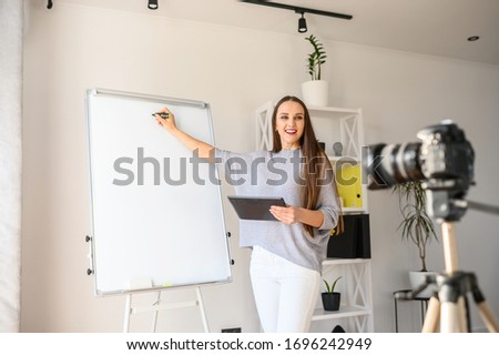 Female teacher, tutor in casual wear makes tutorial video on the camera on a tripod indoors . She with tablet in hand points by marker on a flipchart. The concept of online education, online learning