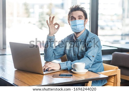 Portrait of positive young man with surgical medical mask in blue shirt sitting and working on laptop and showing okay sign, looking at camera. indoor working and health care concept.