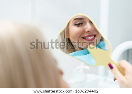 Young woman patient looks at her teeth in the mirror and enjoys a beautiful smile after successful dental treatment or professional oral hygiene. Modern dentistry clinic