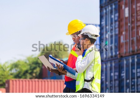 Worker man in hardhat and safety vest holding laptop and Female foreman talks on two-way radio control loading containers box from cargo