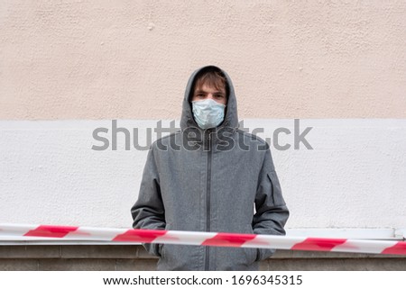 A man stands near the wall in the hood on the street. On face, a medical white mask, protection from coronavirus and viruses transmitted by the drip. Front hangs red white prohibiting tape Royalty-Free Stock Photo #1696345315