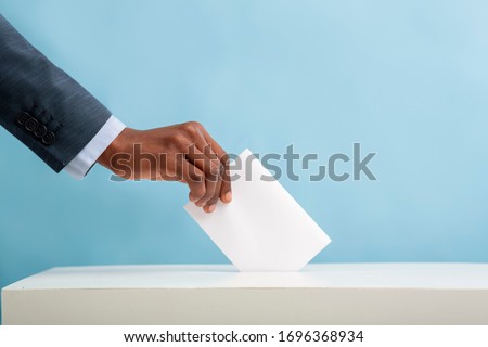 Elections in USA 2020. African american man putting an empty ballot in election box over blue background, copy space Royalty-Free Stock Photo #1696368934