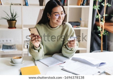 Image of a beautiful happy optimistic young girl student indoors studying using mobile phone holding credit card. Royalty-Free Stock Photo #1696378138