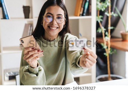 Image of a beautiful cheerful young girl student indoors studying using mobile phone holding credit card.