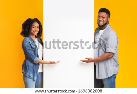 Positive African Couple Pointing At White Advertisement Board With Open Palms, Standing Together Over Yellow Background, Free Place For Your Text Royalty-Free Stock Photo #1696382008