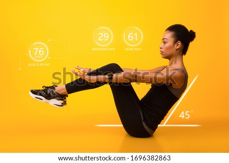 Workout. African american woman doing abs exercise with leg raise over yellow background. Smartwatch shows her body parameters. Studio shot