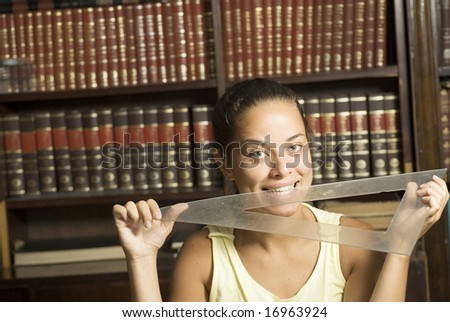 Woman playfully biting on a protractor with books in the background. Horizontally framed photo